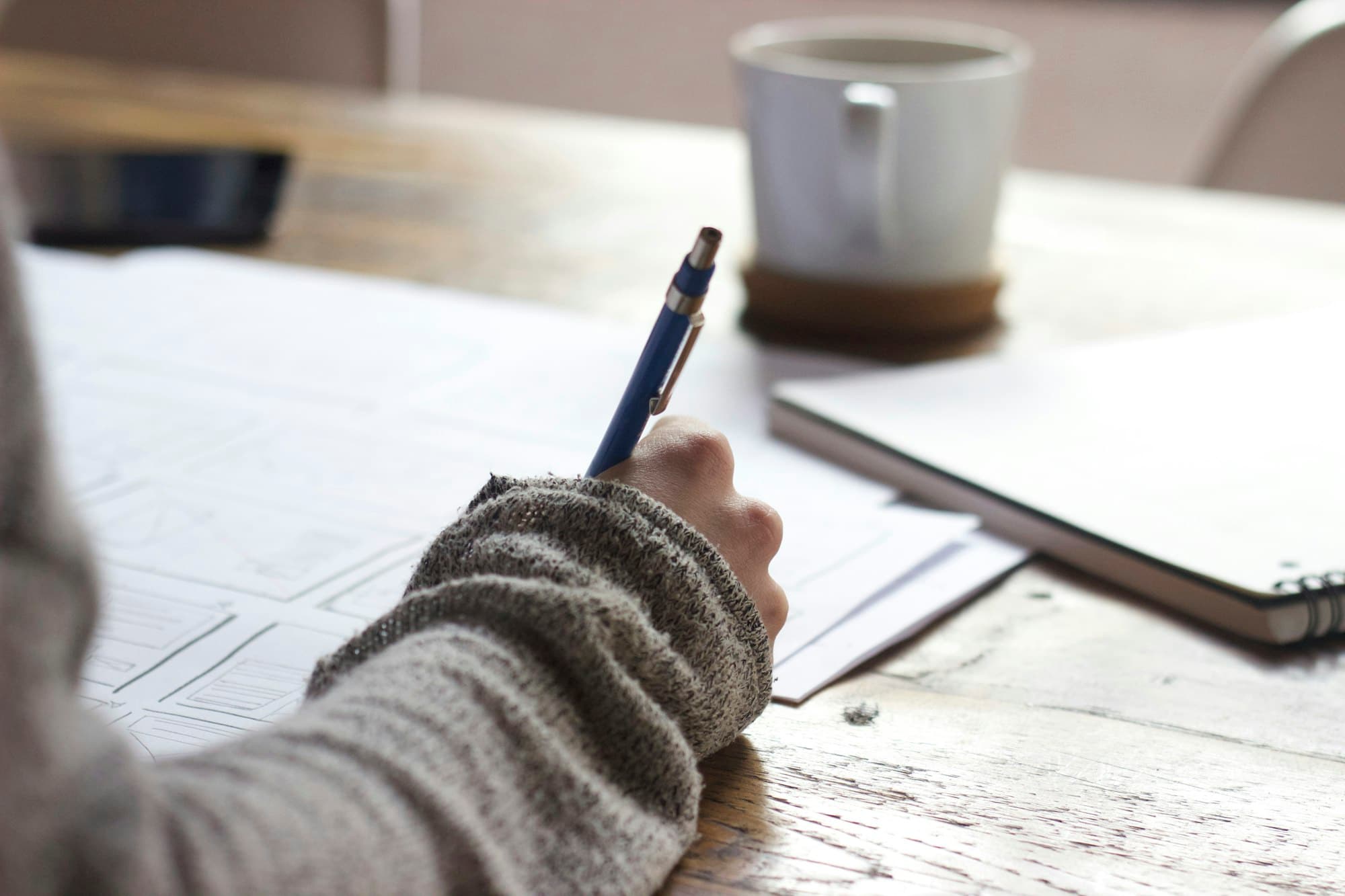 A hand writing notes at a wooden table beside a coffee mug